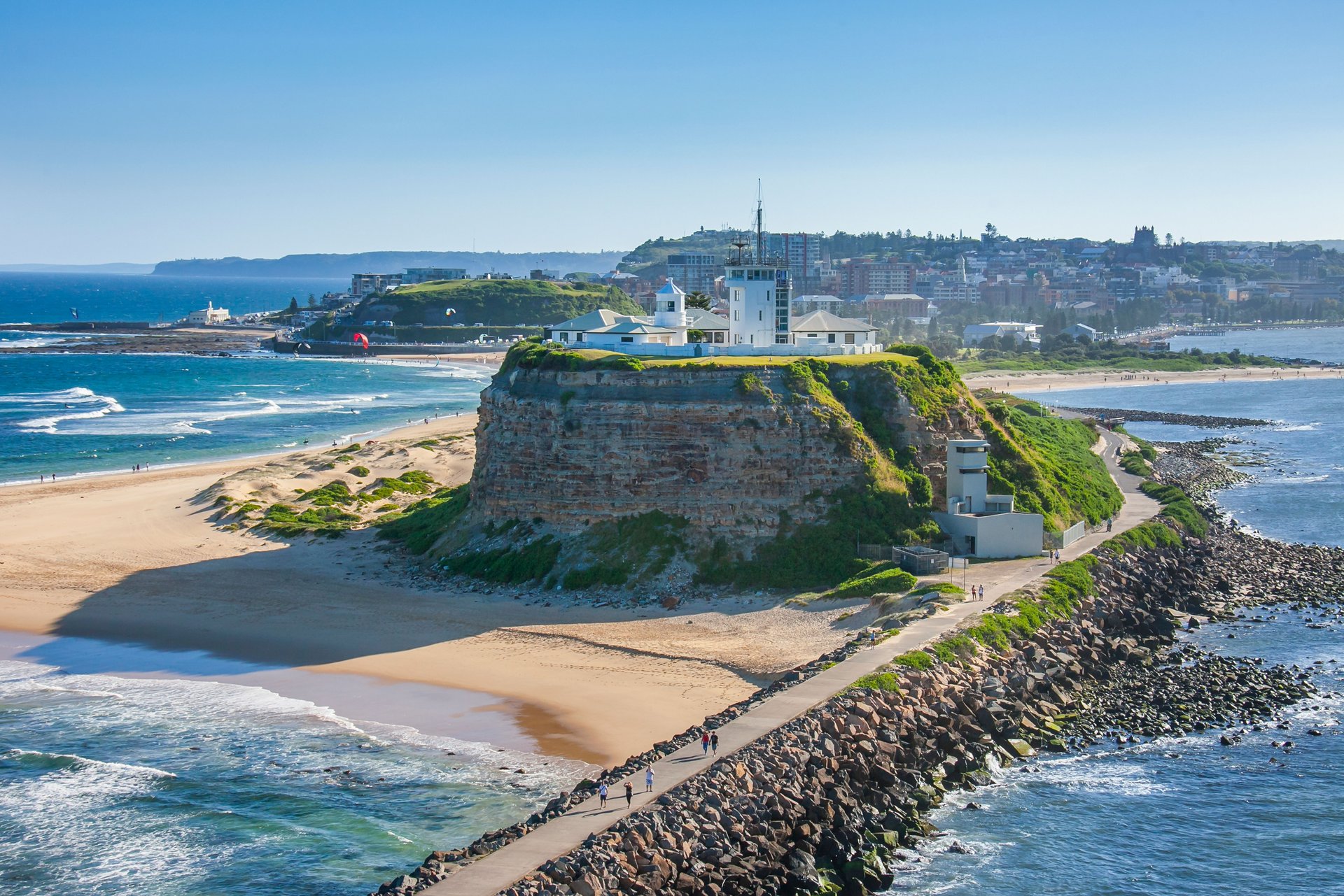 Aerial view of a coastal headland with a lighthouse, surrounded by sandy beaches and ocean waves under a clear blue sky.