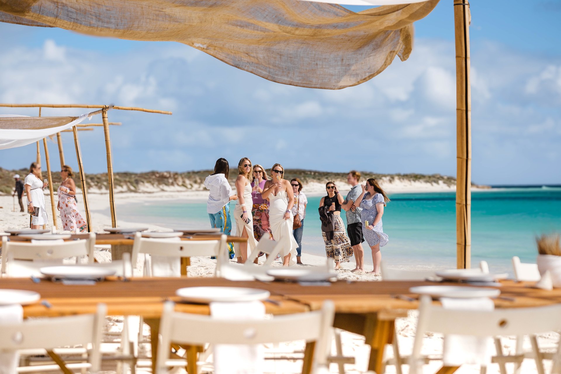 Wedding guests mingling on a white-sand beach by turquoise water beneath a cloth canopy, with wooden tables and chairs set in the foreground.