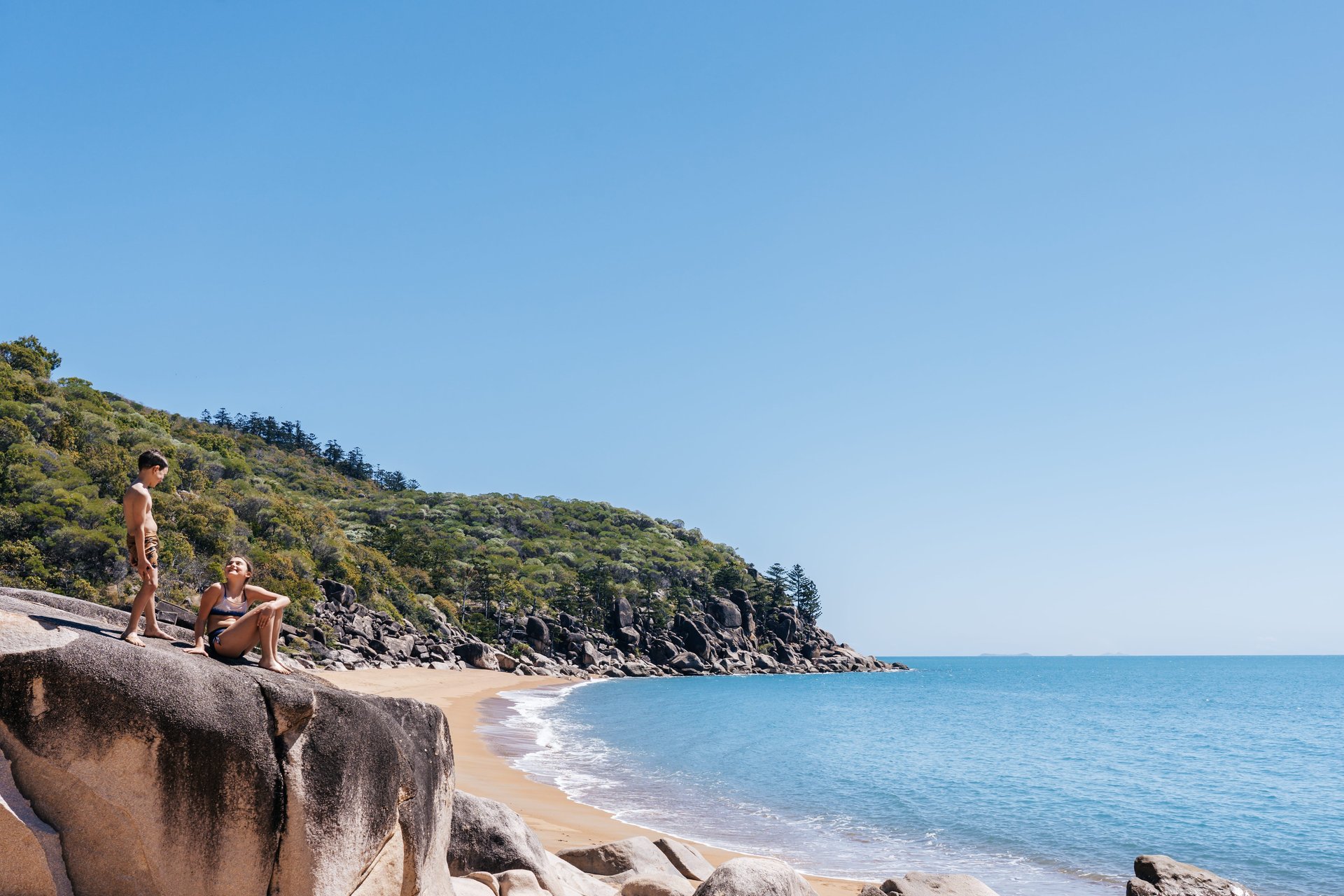 Two people in swimsuits on large coastal rocks overlooking a sandy beach, blue sea and forested headland under a clear sky.