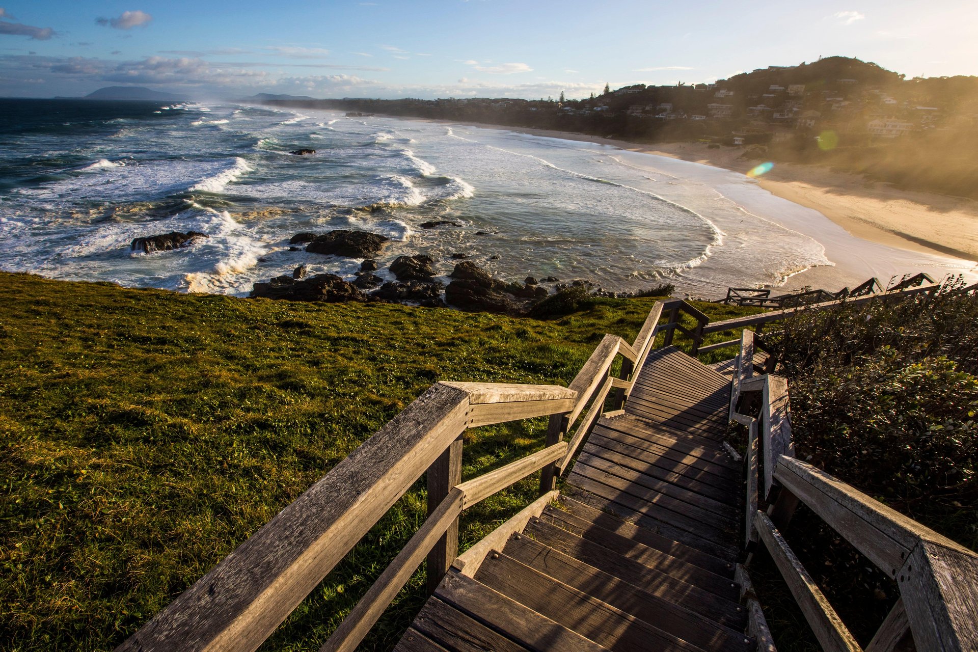 Wooden stairs down a grassy coastal bluff to a sandy beach, waves breaking along the sunlit shoreline.