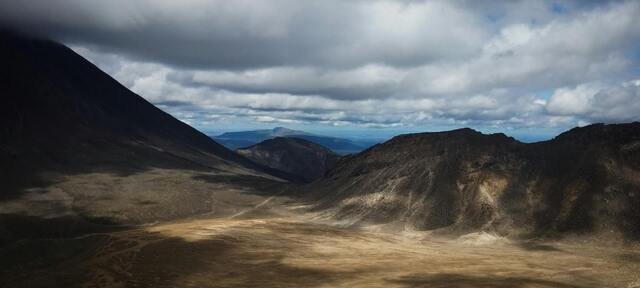 A vast, barren landscape with rugged mountains under a cloudy sky, casting shadows on the dry terrain below.