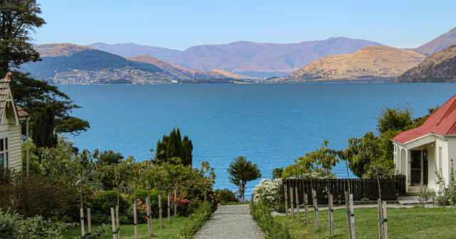 Pathway leading to a lake surrounded by hills, with houses and gardens on either side, under a clear blue sky.