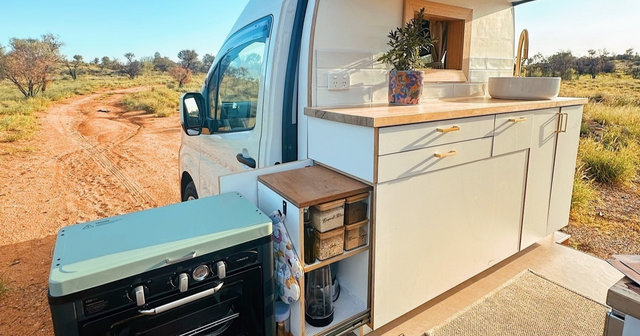 Camper van kitchen setup with open door revealing a countertop, sink, storage shelves, and a portable stove in a desert landscape.