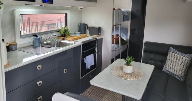 Modern RV interior with a kitchenette, sink, oven, and dining area. Features gray cabinets, a small plant, and bunk beds in the background.