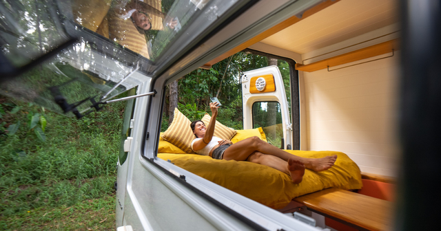 Woman relaxing on a yellow bed inside a camper van, taking a selfie. A man smiles through an open window. Lush greenery outside.