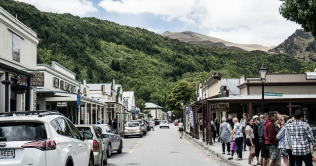 A bustling street in a small town with parked cars, shops, and a crowd of people, set against a backdrop of lush green hills.