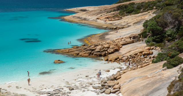 A scenic beach with turquoise water, rocky shoreline, and a few people enjoying the sun and sea. Lush greenery borders the sandy area.