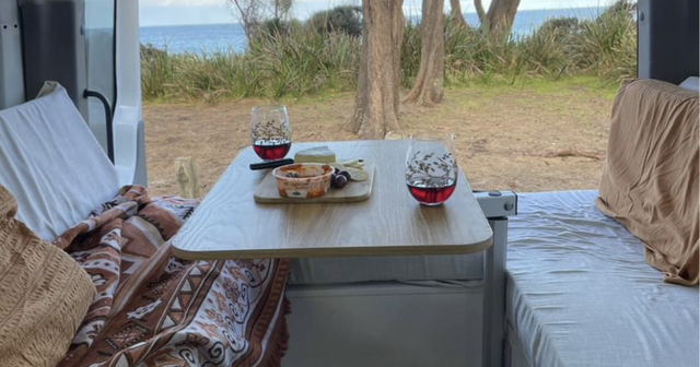 Interior of a camper van with a wooden table set for two, featuring wine, cheese, and snacks, overlooking a beach view with trees.
