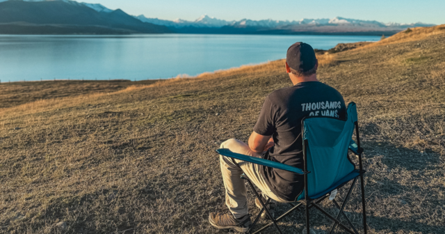 A person sits in a camping chair on a grassy hill, overlooking a lake and distant snow-capped mountains under a clear blue sky.
