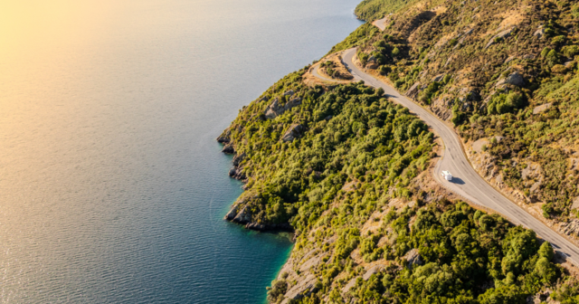 Aerial view of a winding coastal road with a vehicle, surrounded by lush greenery and adjacent to a calm blue sea under a warm sky.