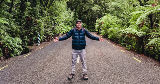 Person in a puffy jacket stands with arms open on a forest road, surrounded by lush green trees and ferns.
