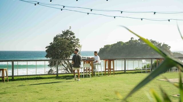 Two people at a grassy outdoor area with tables, overlooking the ocean and a small island. String lights are hung above.