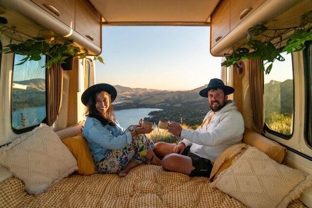 A couple sits in a cozy van interior, toasting with mugs, overlooking a scenic lake and mountain view through the open back doors.