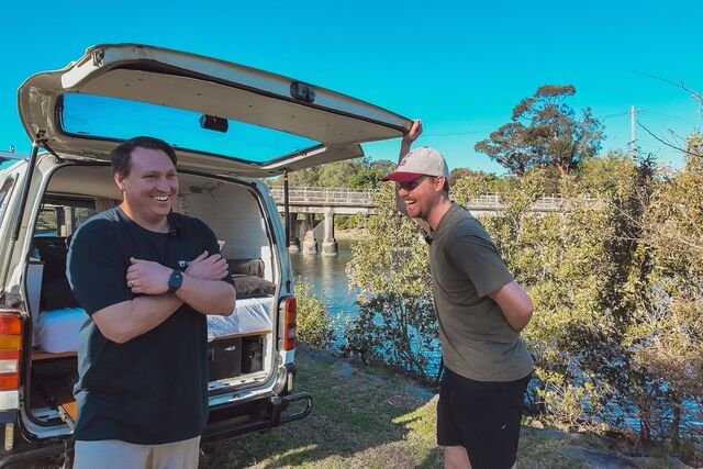 Two men laughing near an open van by a river, with a bridge and trees in the background under a clear blue sky.