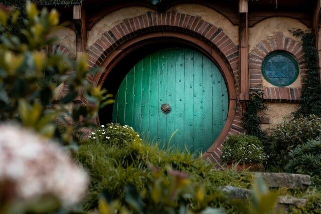 A round, green wooden door set in a brick archway, surrounded by lush greenery and small flowers, evoking a whimsical, rustic atmosphere.