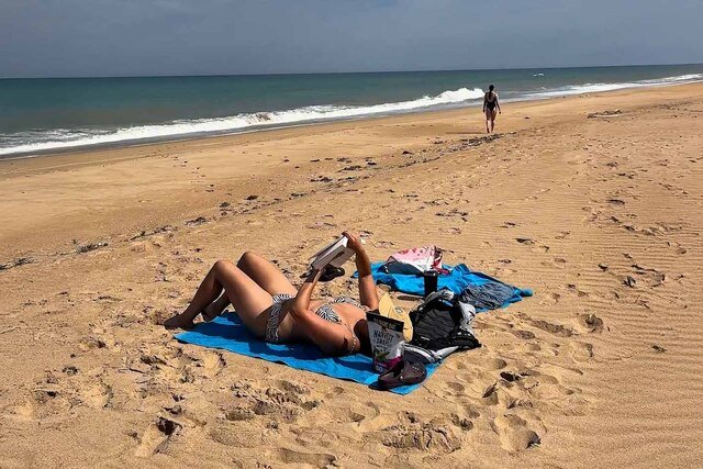 A woman laying on a beach towel at the beach reading a book