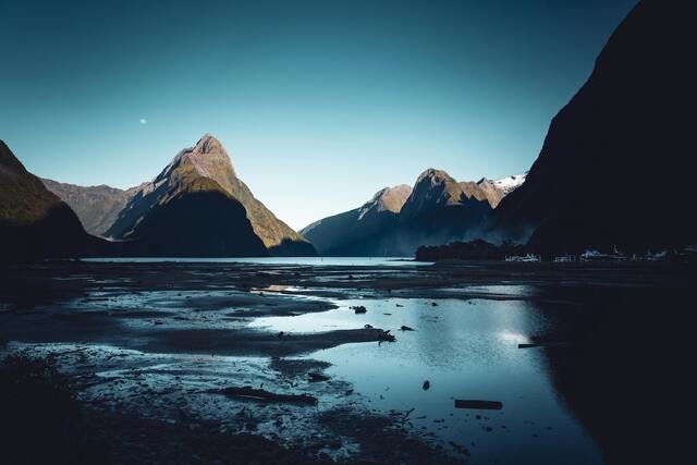 A serene fjord landscape at dusk, with towering mountains, calm water reflecting the sky, and a small moon visible.