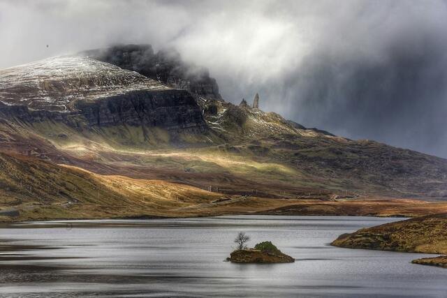 A serene lake with a small island in the foreground, surrounded by misty mountains and a cloudy sky. Snow dusts the mountain peaks.