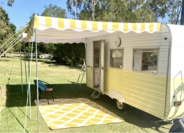 A vintage yellow and white camper with a striped awning, set up on grass with a matching rug and chair outside. Trees are in the background.