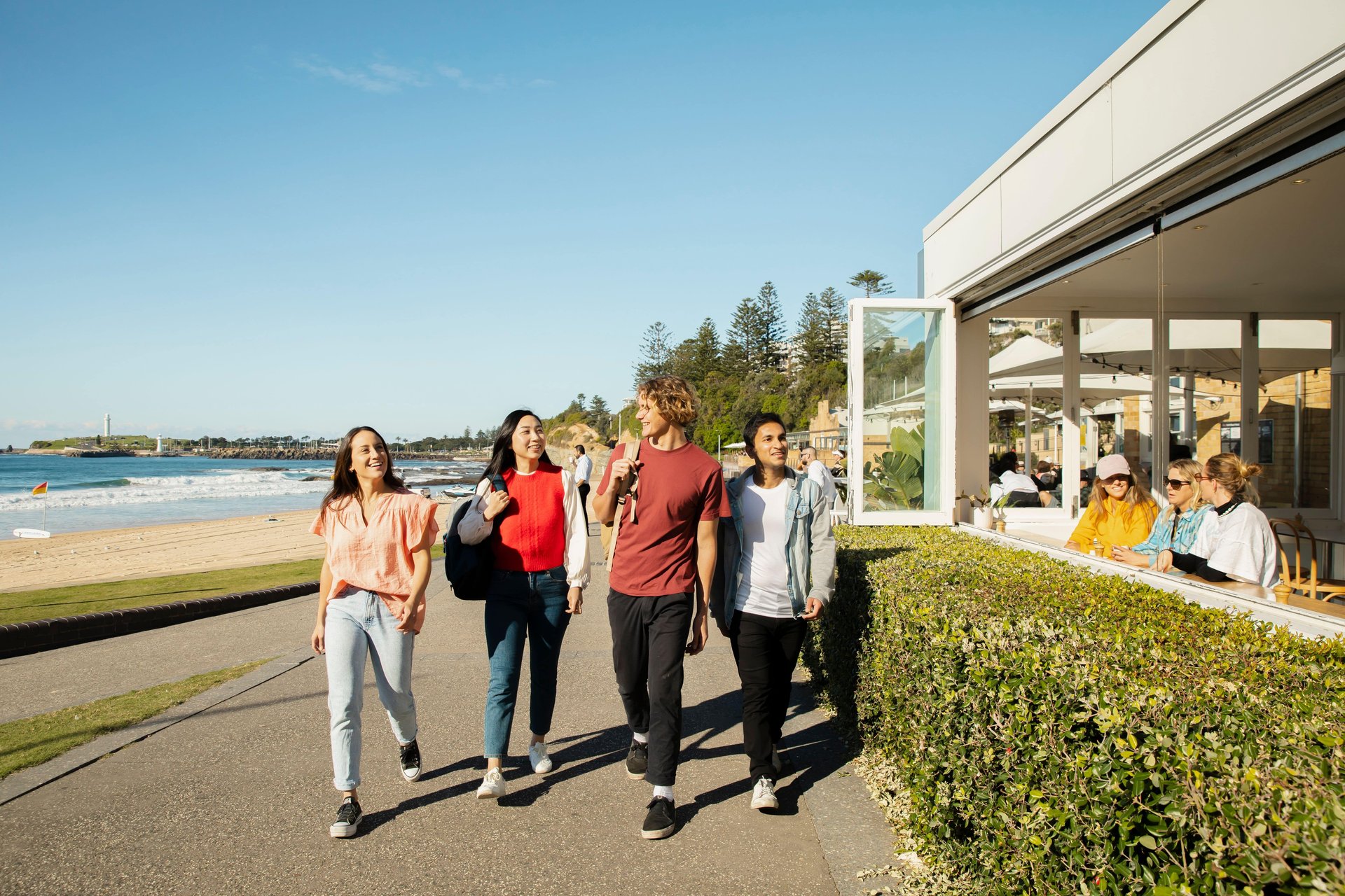 A group of 4 friends walk along a beachside path near a cafe