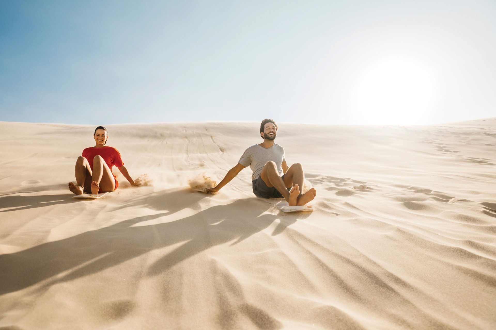 Two people sliding down a sunlit sand dune, smiling and enjoying a bright, clear day.