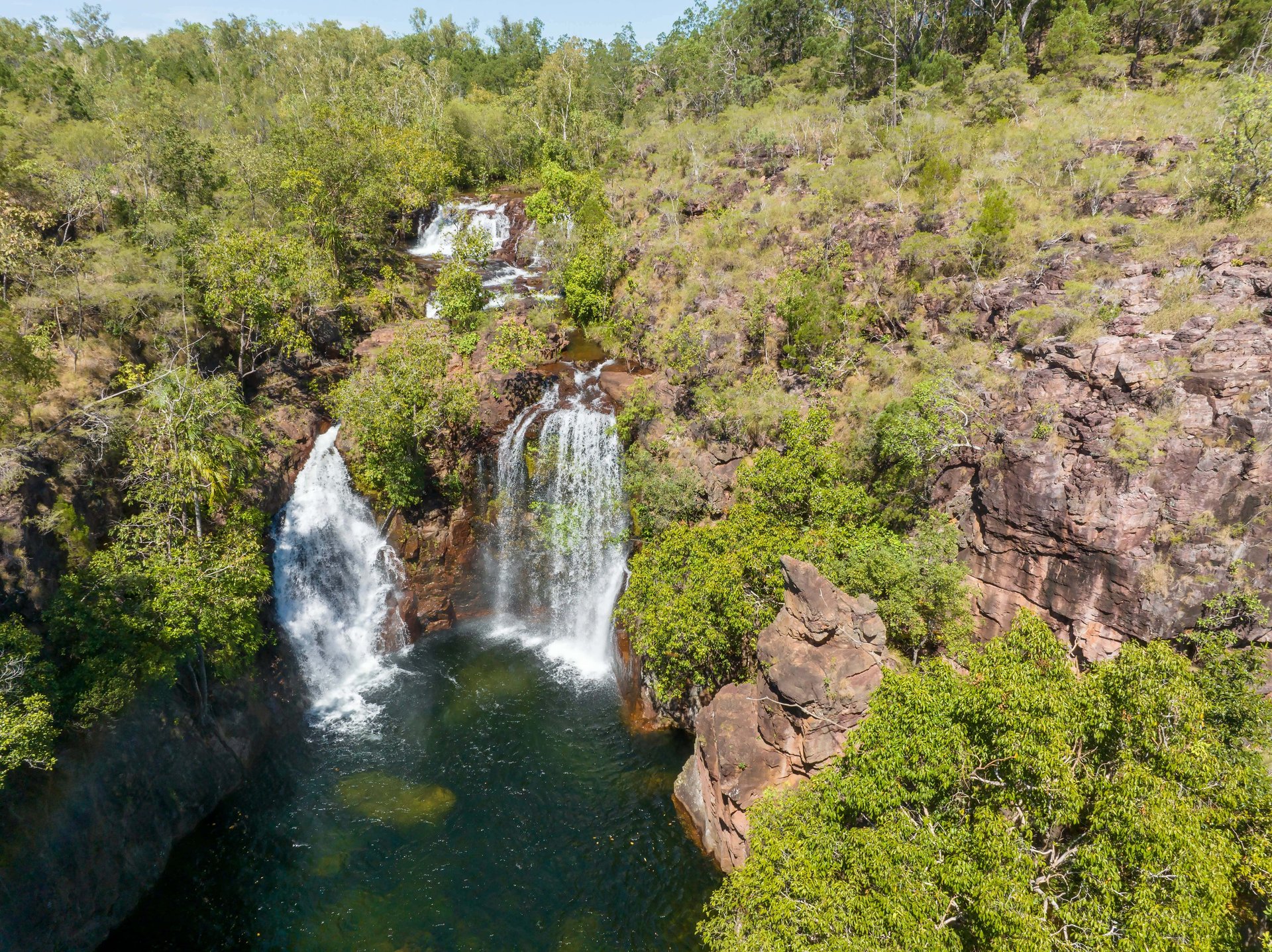 Aerial view of twin waterfalls cascading into a deep green pool surrounded by red-brown rocky cliffs and lush trees.