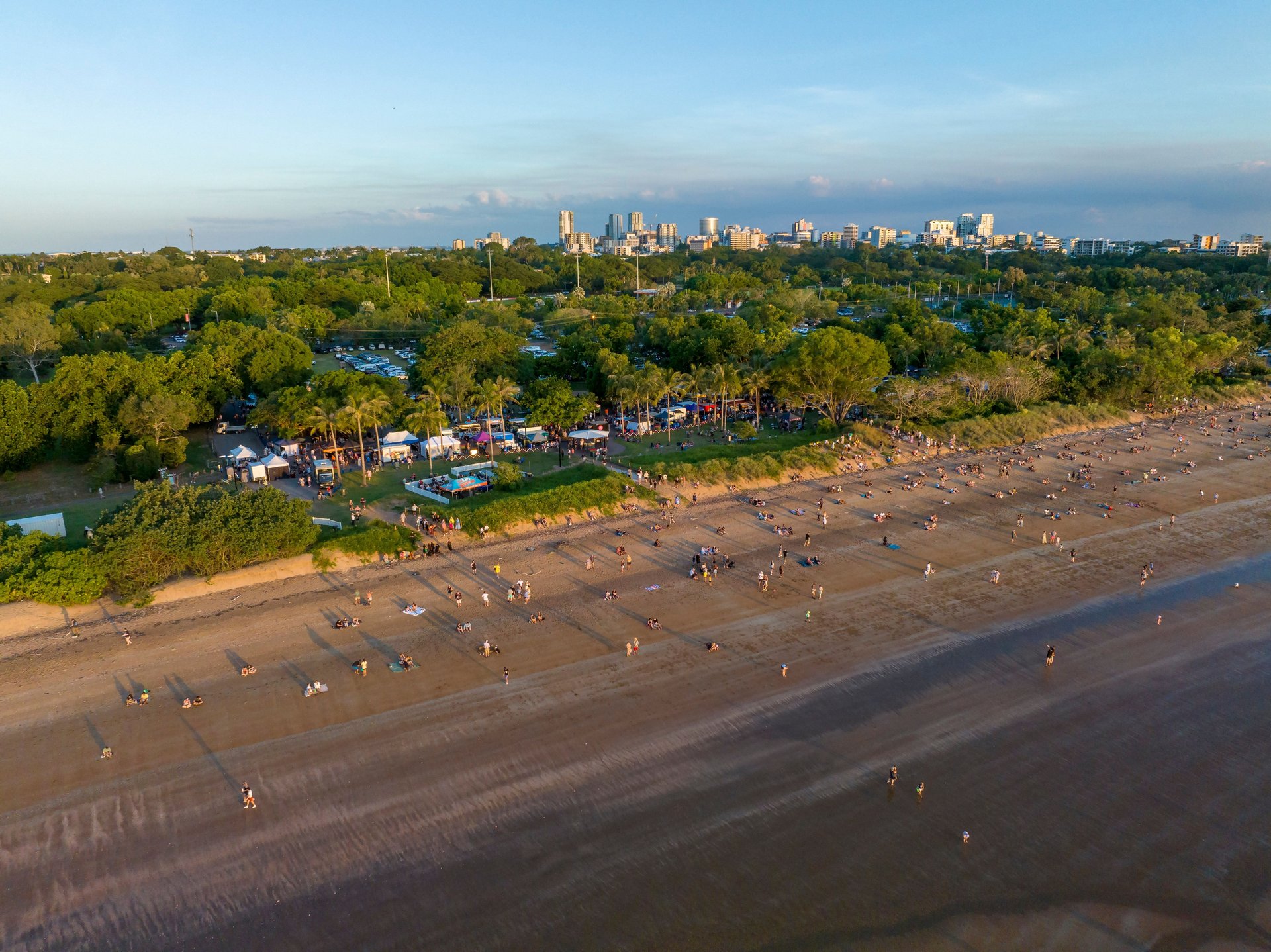Aerial view of a sandy beach with scattered people, grassy park and festival tents behind it, green trees and a distant city skyline at sunset.