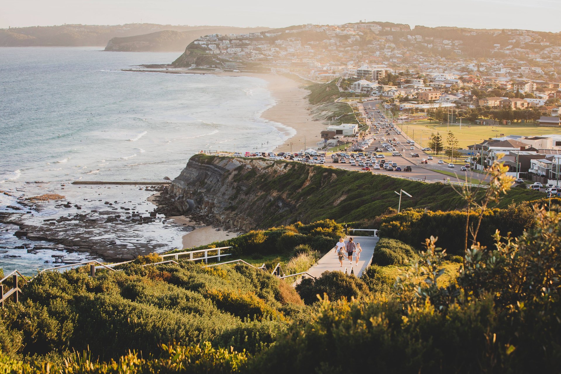 Coastal view with a winding road, cliffs, and a sandy beach. People walk along a path surrounded by greenery, with a town in the background.