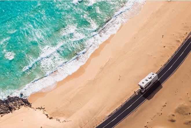 Vista aérea de una caravana blanca conduciendo por una carretera costera junto a una playa de arena y olas del océano turquesa.