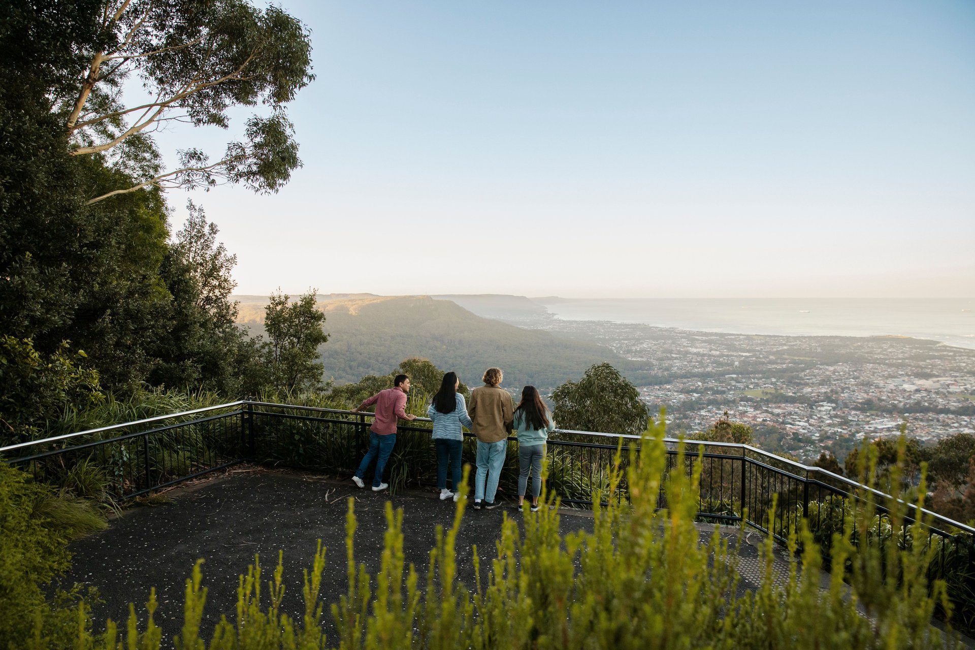 Four people at a lookout railing viewing a forested valley, town and distant coastline.