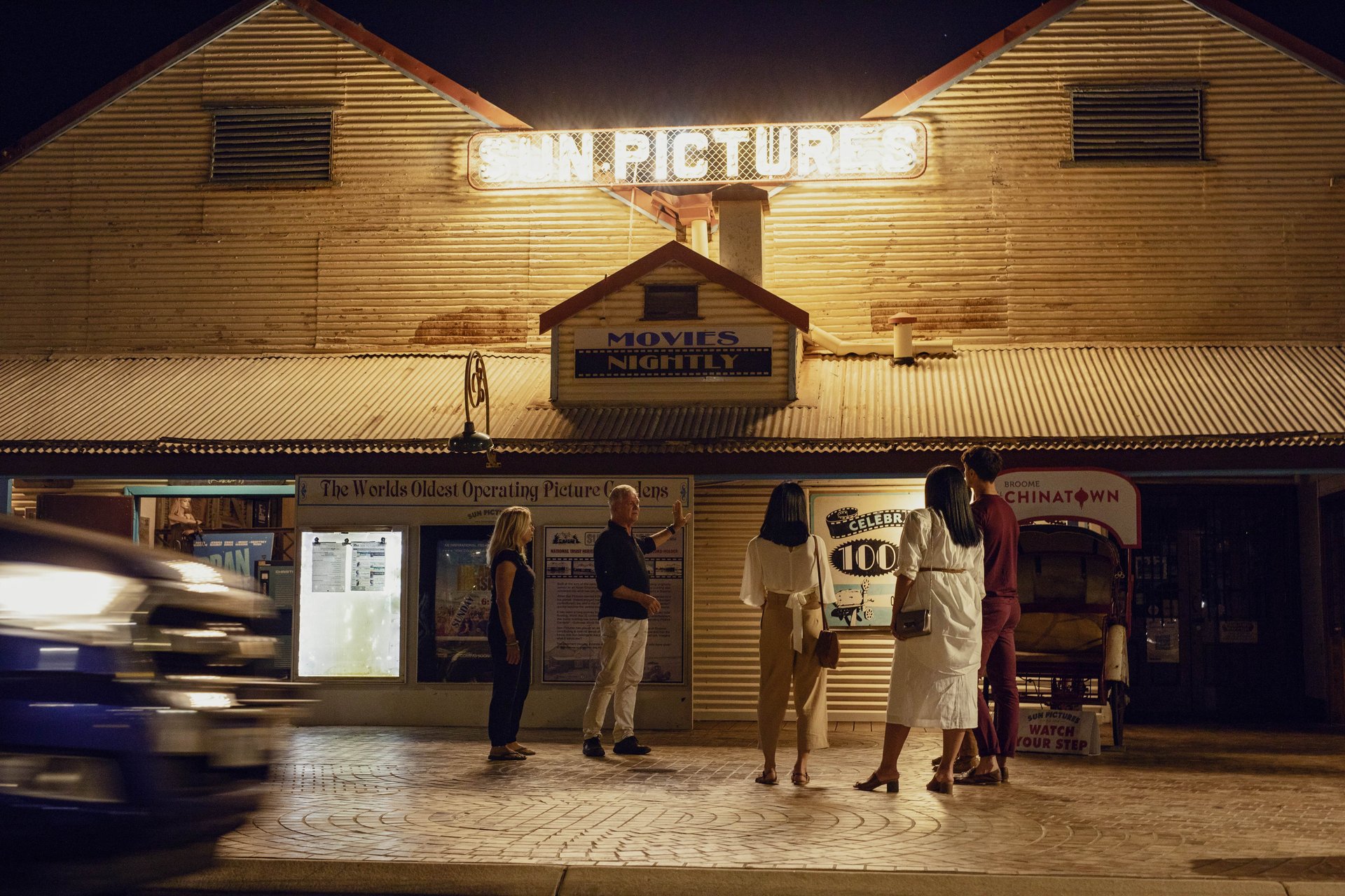 Night view of Sun Pictures theater with illuminated marquee; five people reading posters outside as a car blurs past.