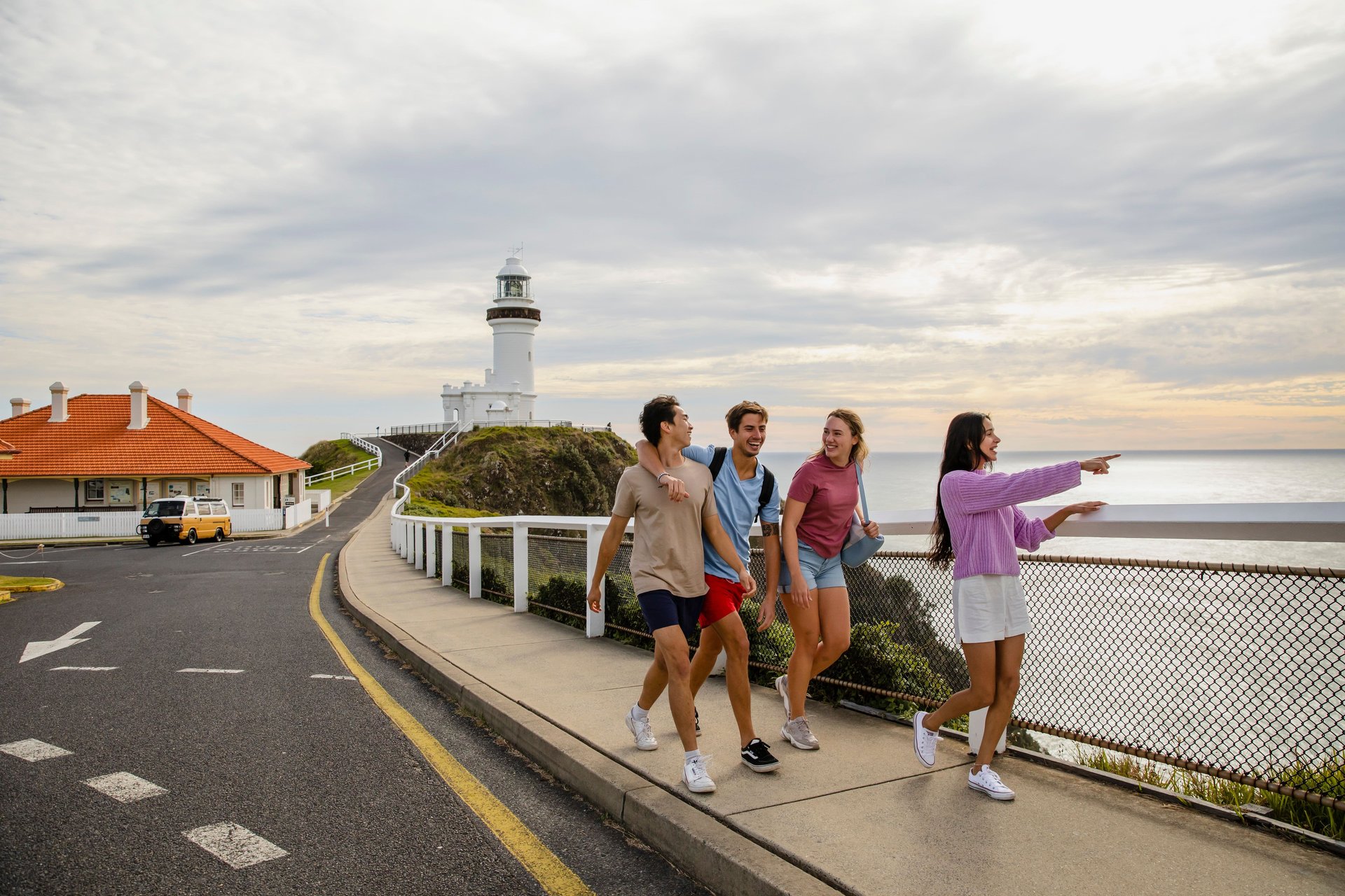 Four friends walking along a seaside path by a white lighthouse, one pointing toward the ocean under a cloudy sky.