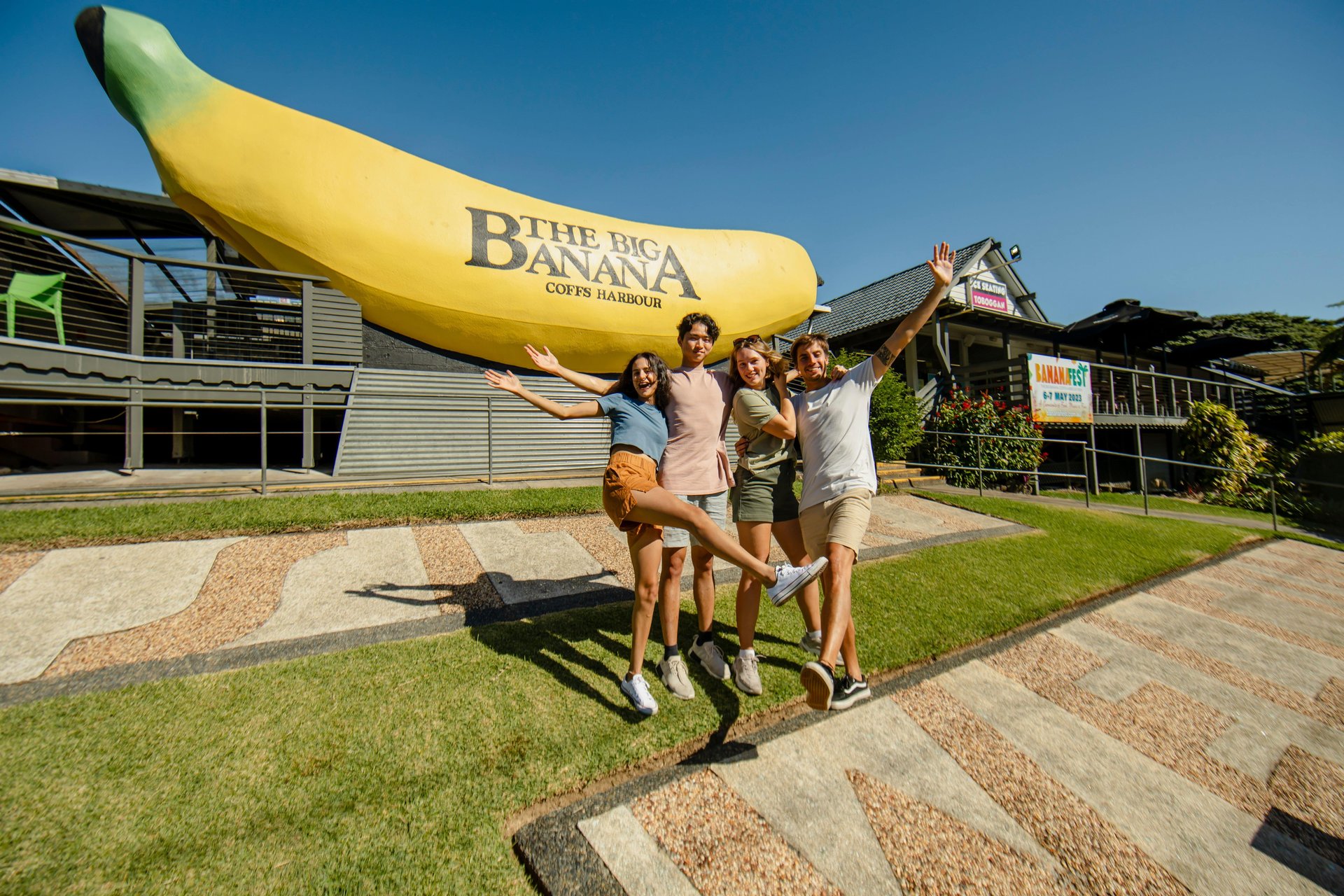 Four friends laughing and posing in front of the giant "The Big Banana" sculpture at Coffs Harbour on a sunny day.