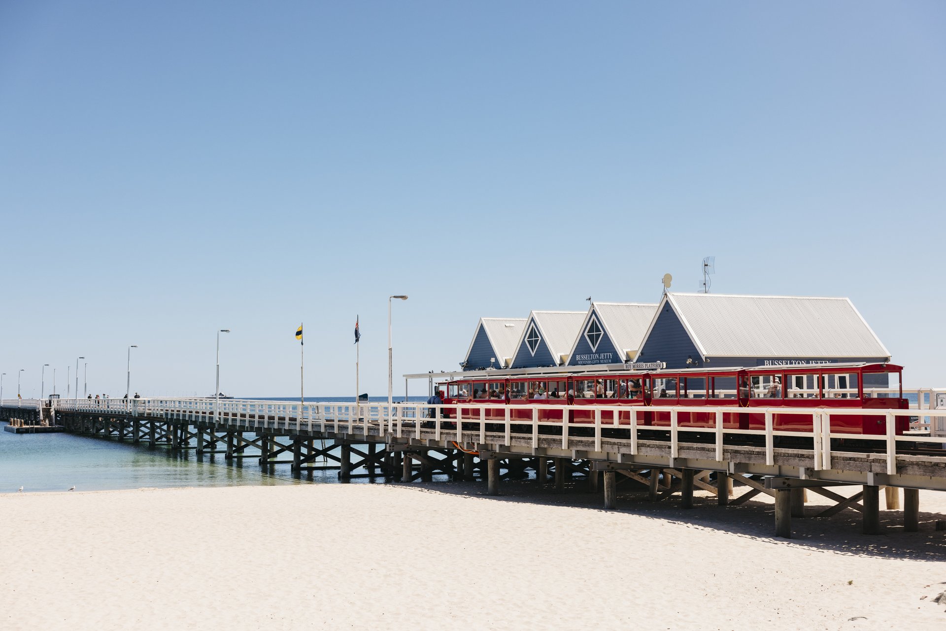 A long wooden pier with small buildings extends over turquoise water, under a clear blue sky. Flags are visible along the pier.