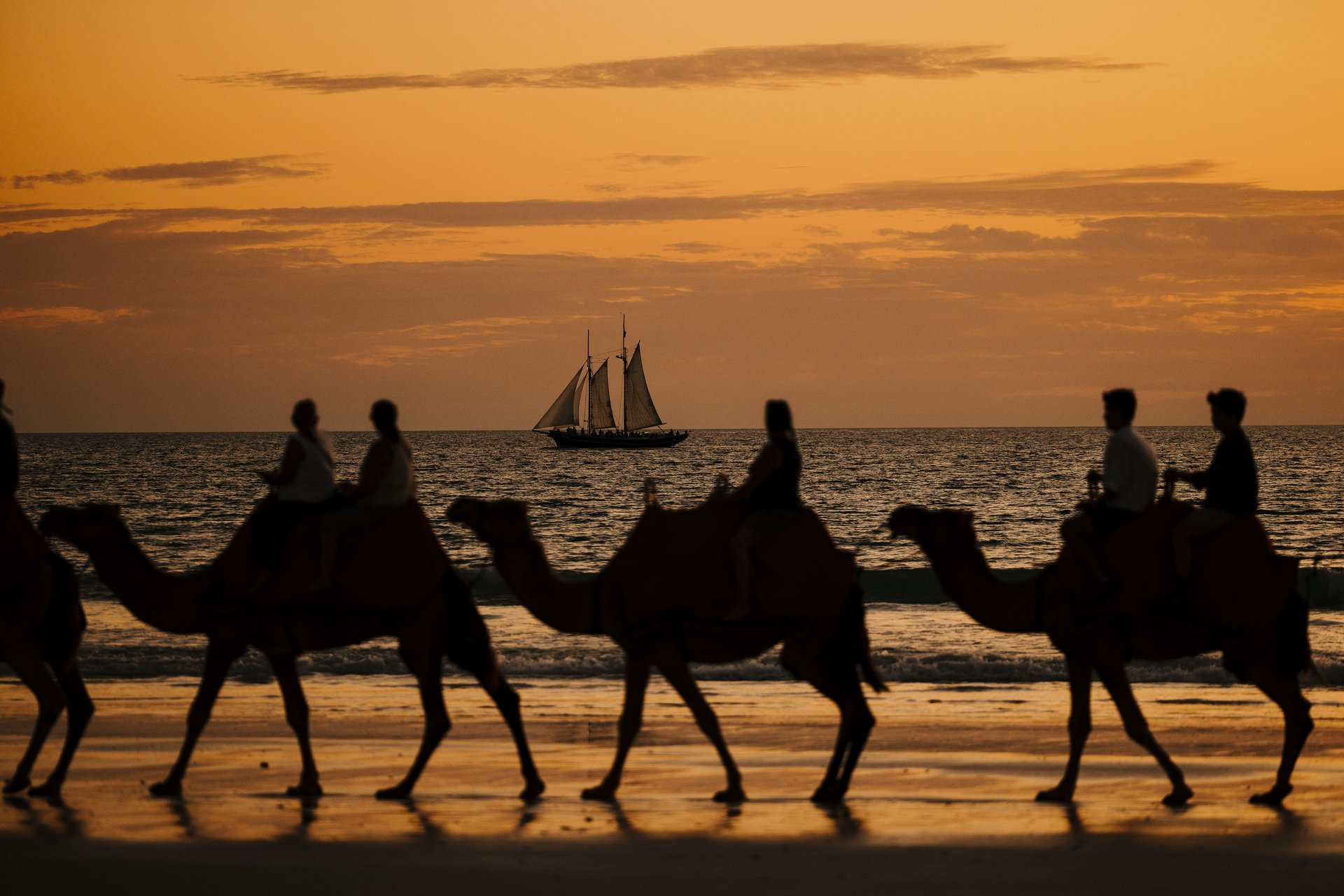 Silhouettes of people riding camels along a beach at sunset, with a sailboat on the horizon under an orange sky.