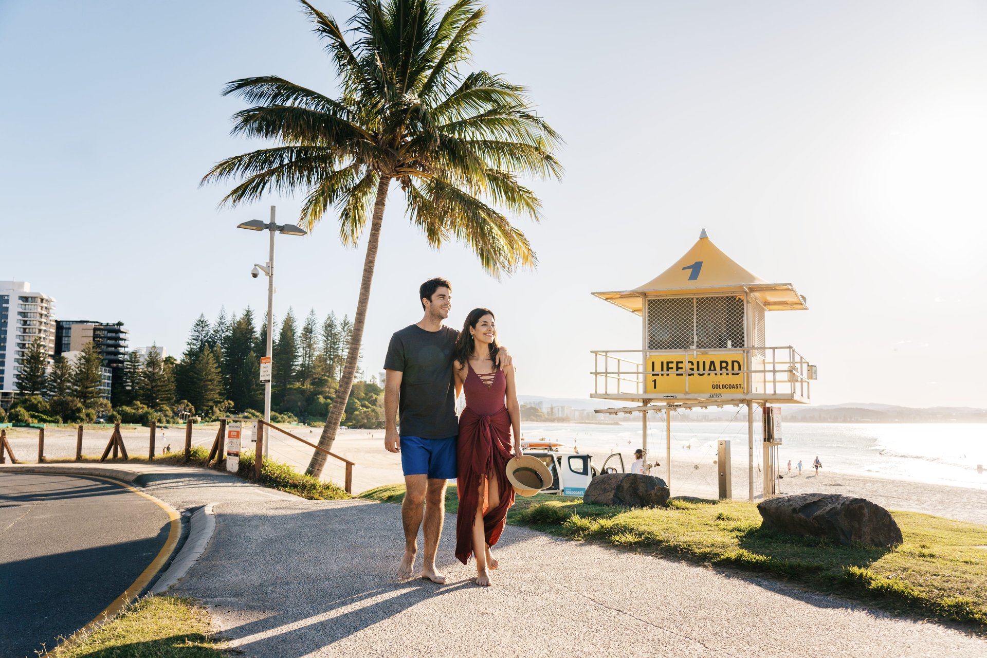 A couple walks barefoot on a beach path near a lifeguard tower, with a palm tree and ocean in the background under a clear sky.