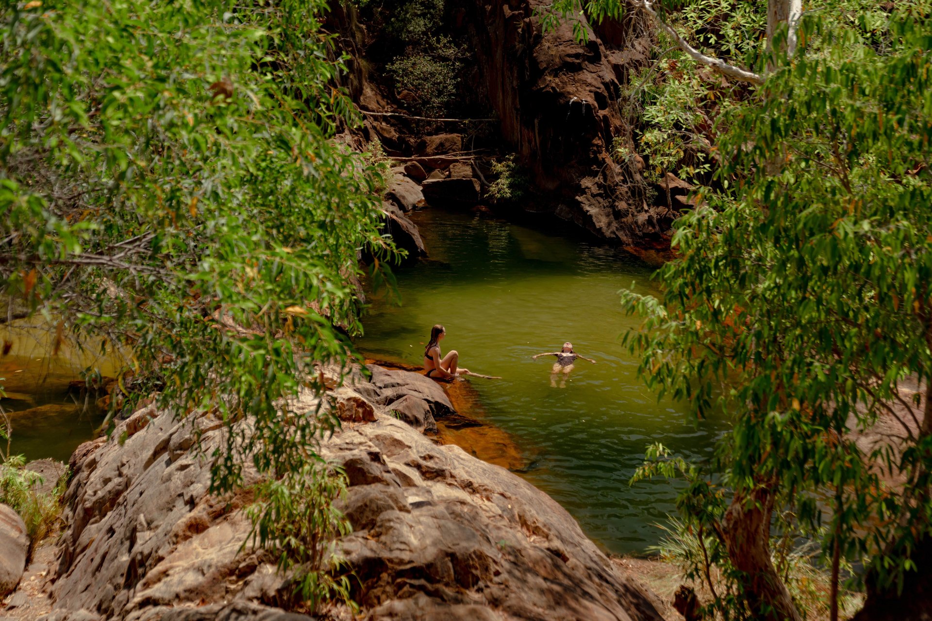 Two people at a shaded rock pool, one sitting on a rock and the other floating on their back in green water, framed by trees.