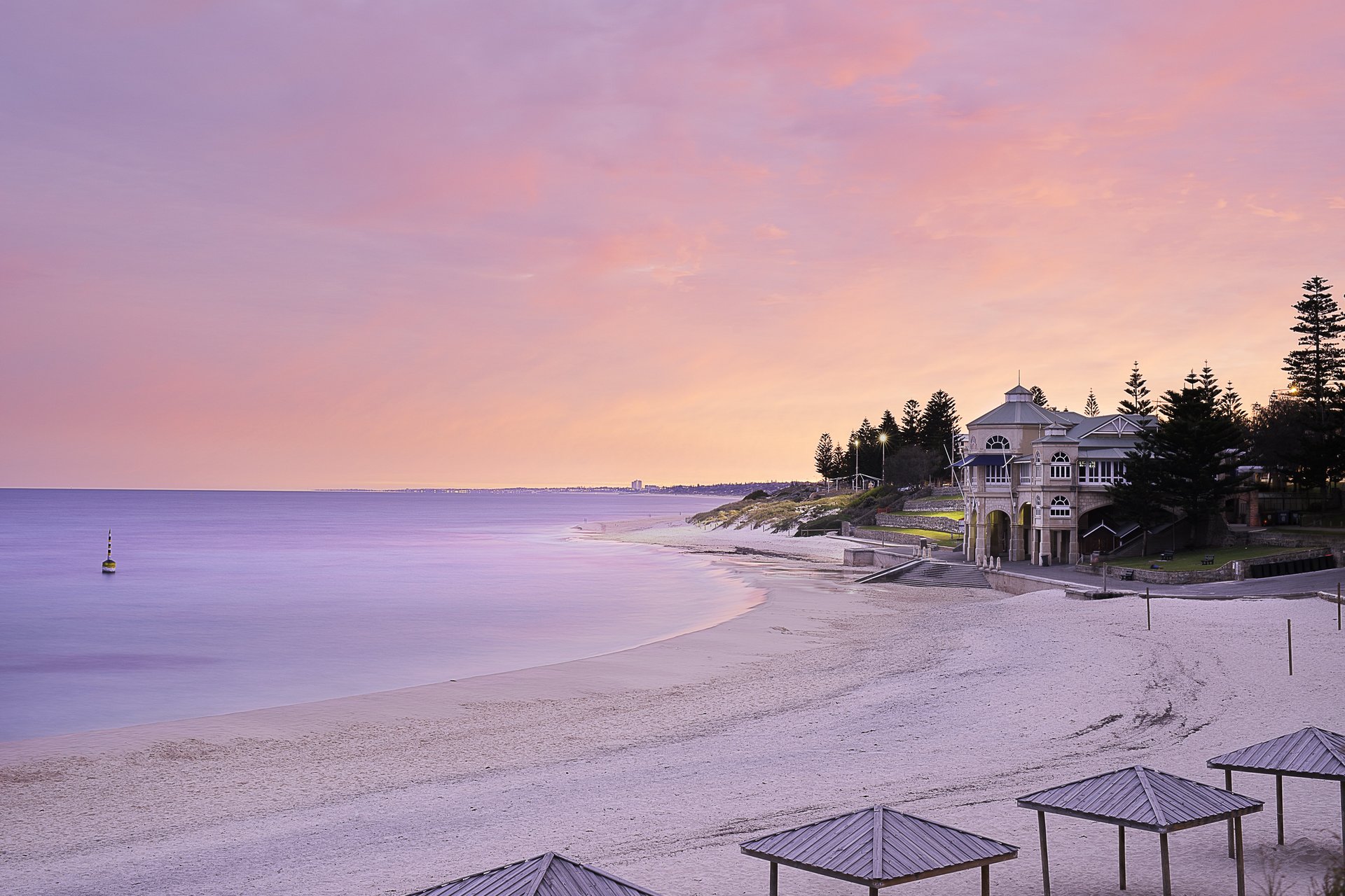 A serene beach at sunset with pastel skies, calm waters, and a historic building surrounded by trees on the shore.