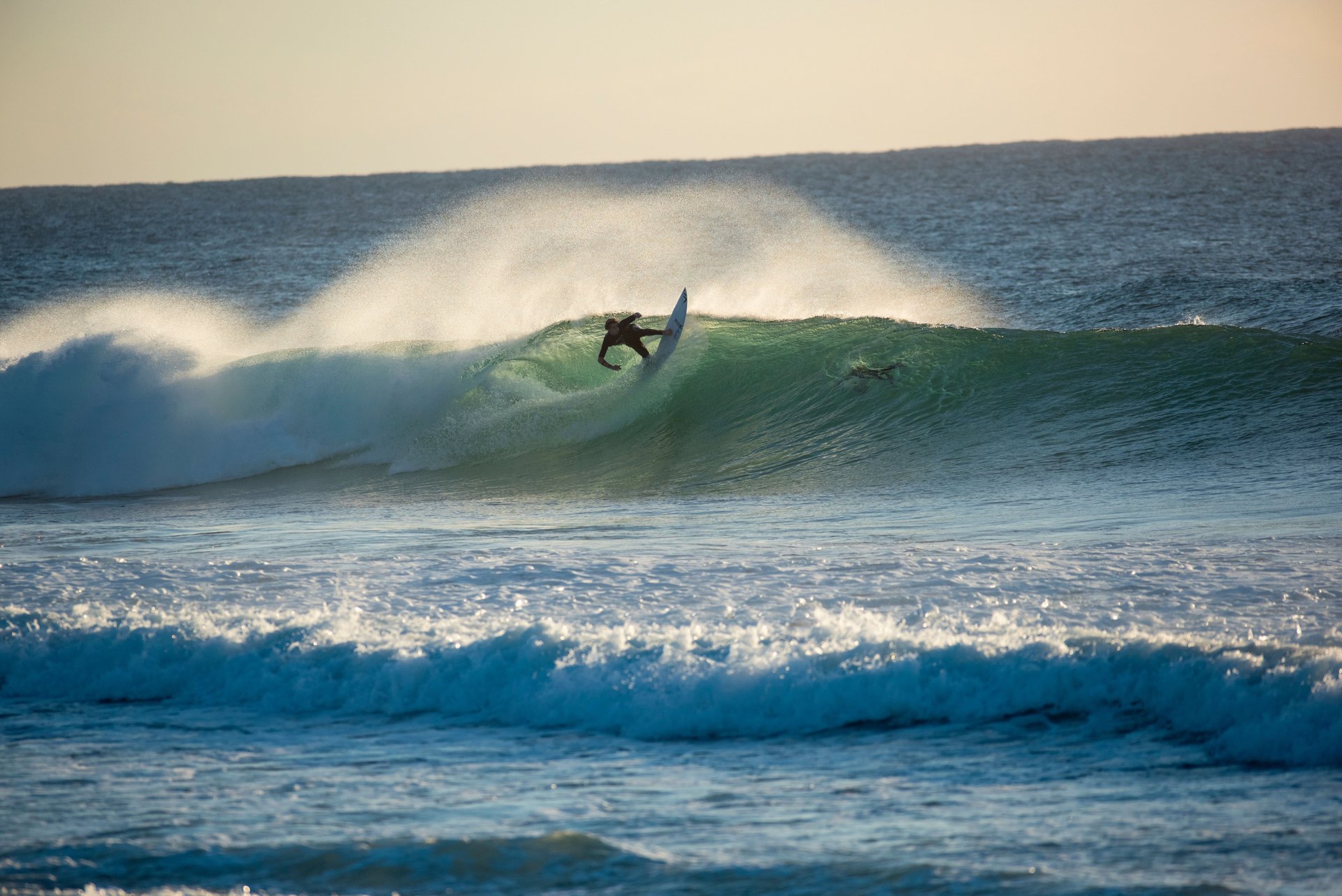 Surfer carving a green wave at sunset, spray arching behind the board.