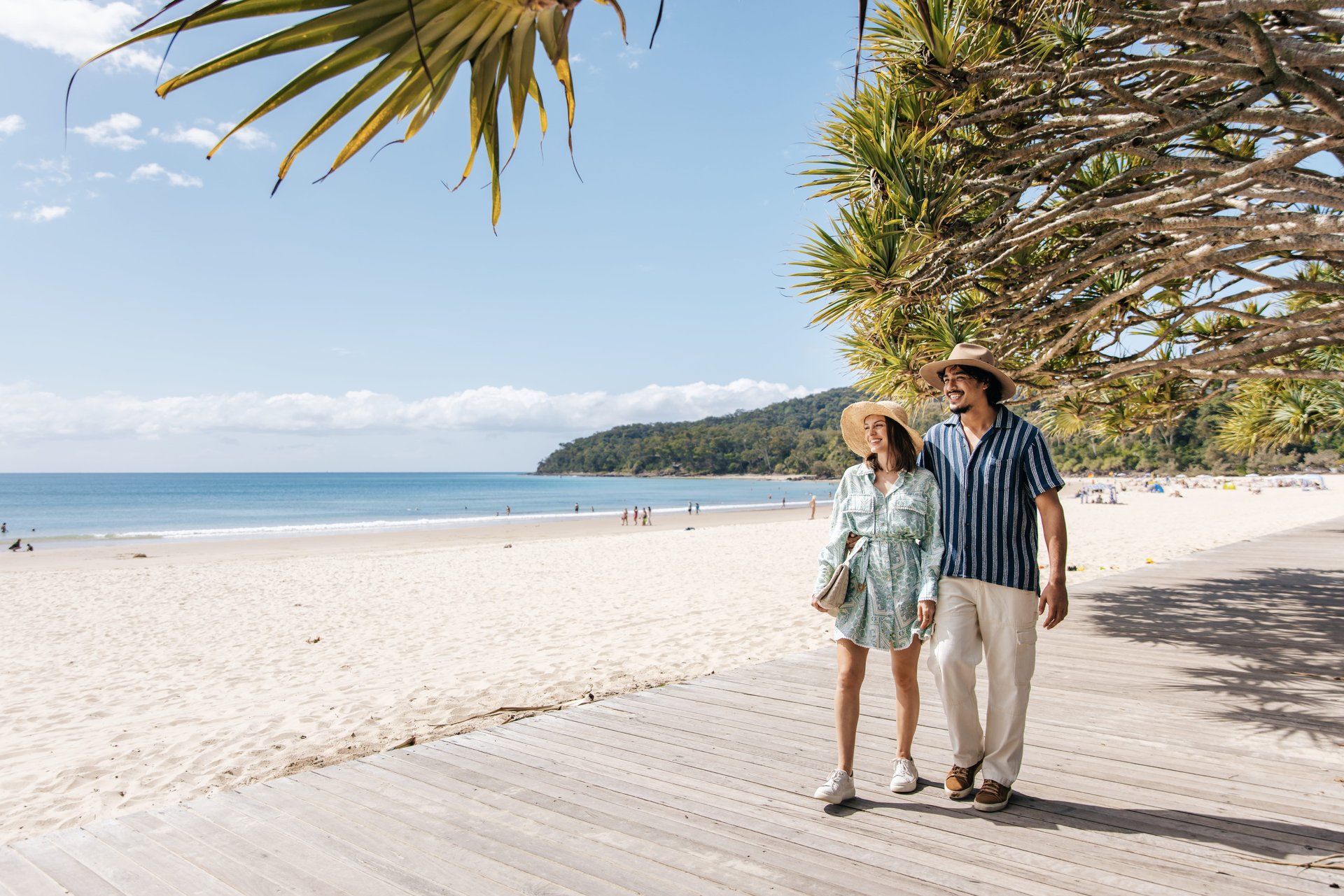 A couple wearing hats walks along a sunny beachside boardwalk, with clear blue skies, sandy beach, and ocean waves in the background.