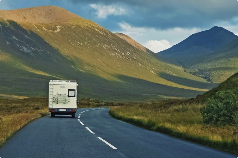 A camper van drives along a winding road through a scenic valley with green hills and mountains under a cloudy sky.