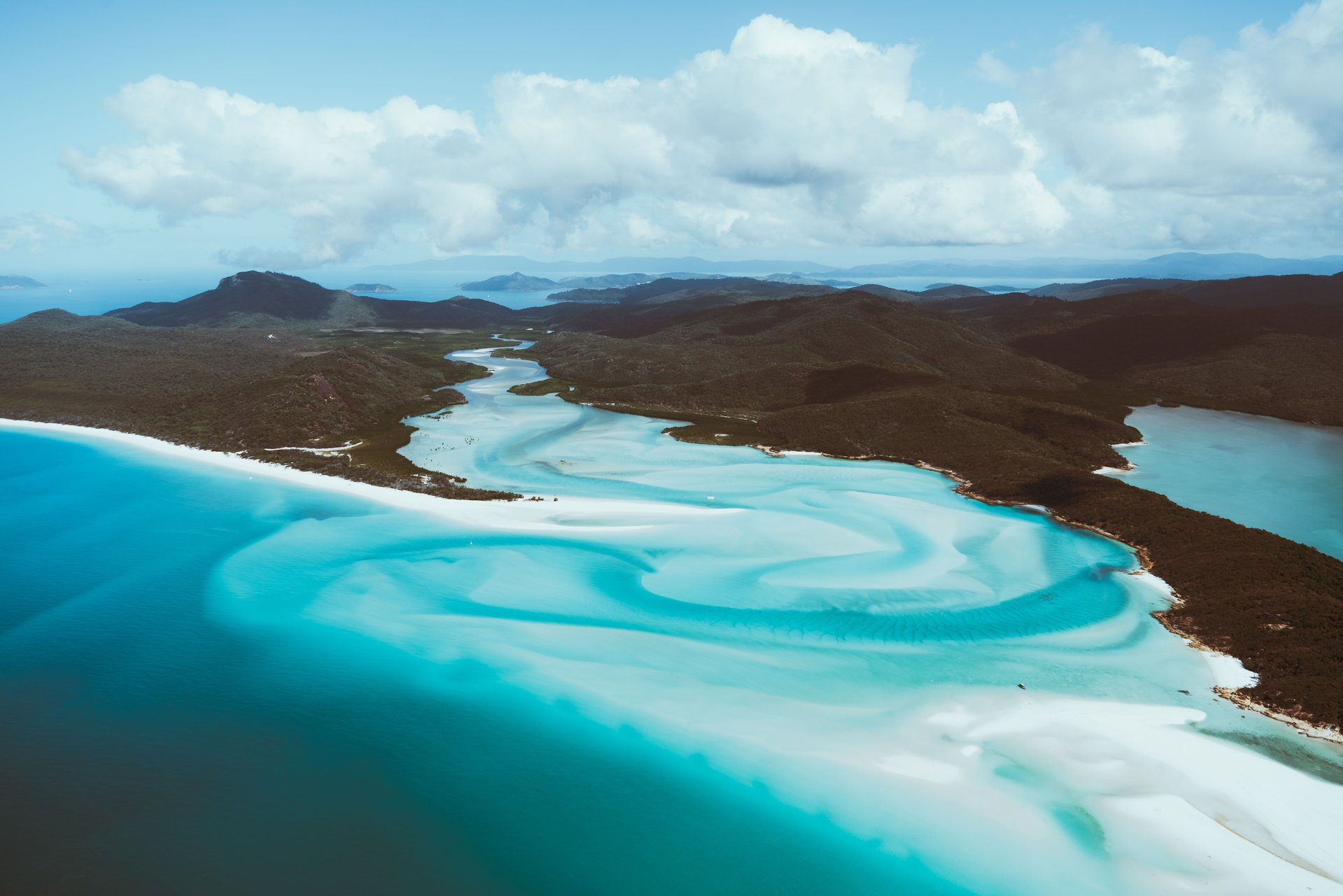 Aerial view of Whitsunday Islands' turquoise waters and swirling white sands under a partly cloudy sky. Lush green hills surround the bay.