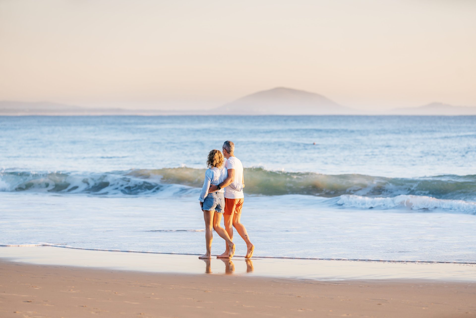 A couple walks along a sandy beach, holding hands, with gentle waves and a distant mountain under a soft, pastel-colored sky.