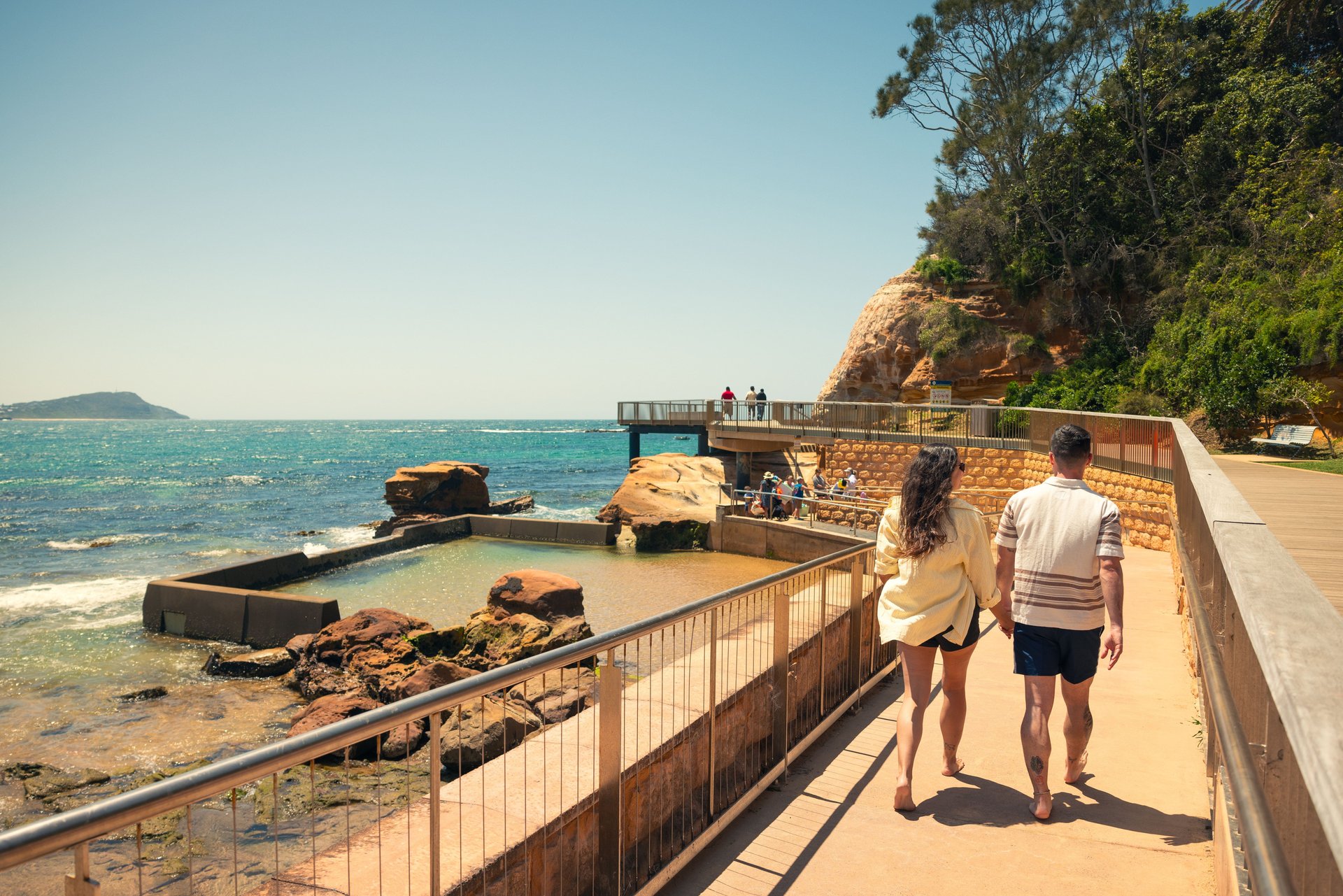Couple holding hands walking barefoot on a sunny coastal boardwalk beside a rock pool and turquoise sea with cliffs behind.