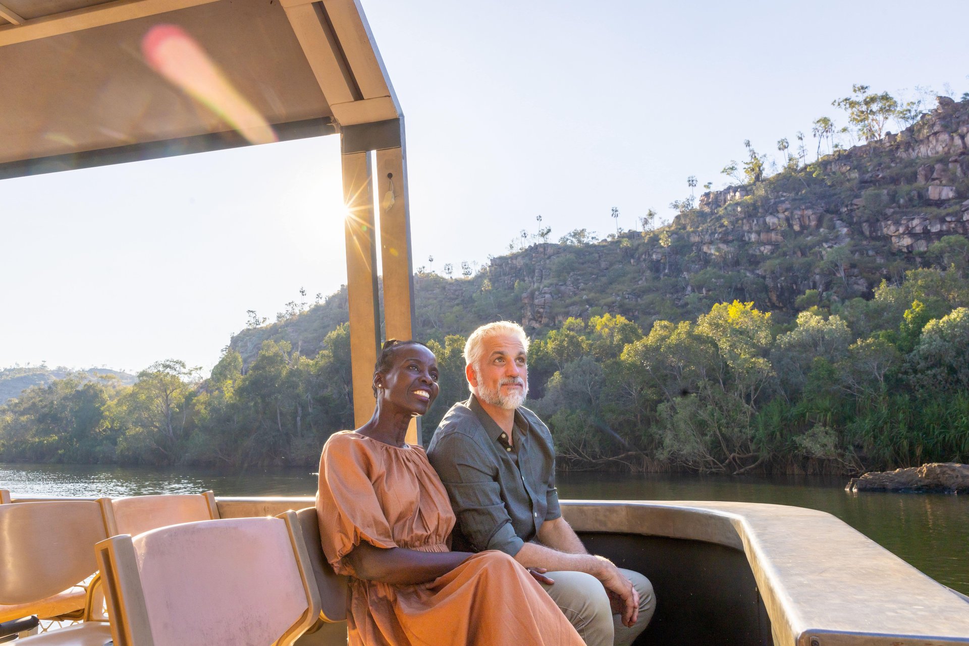 Two people seated on a boat, smiling as sunlight flares, with a calm river and tree-covered rocky hills in the background.