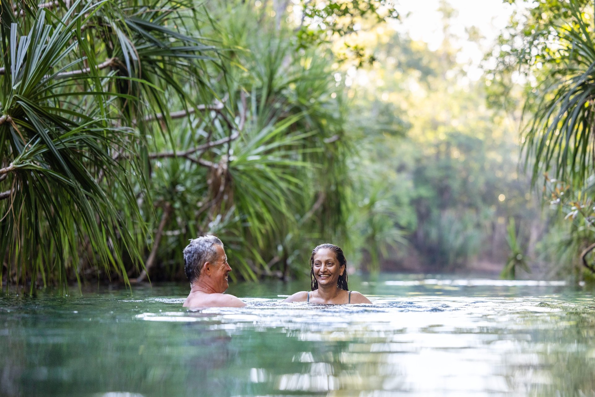 Man and woman smiling while swimming chest-deep in a calm river surrounded by dense tropical foliage