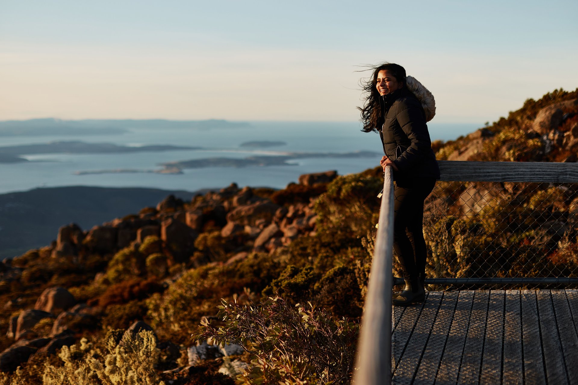 A woman stands on a wooden platform, smiling and leaning on a railing, overlooking a scenic view of rocky hills and distant water.