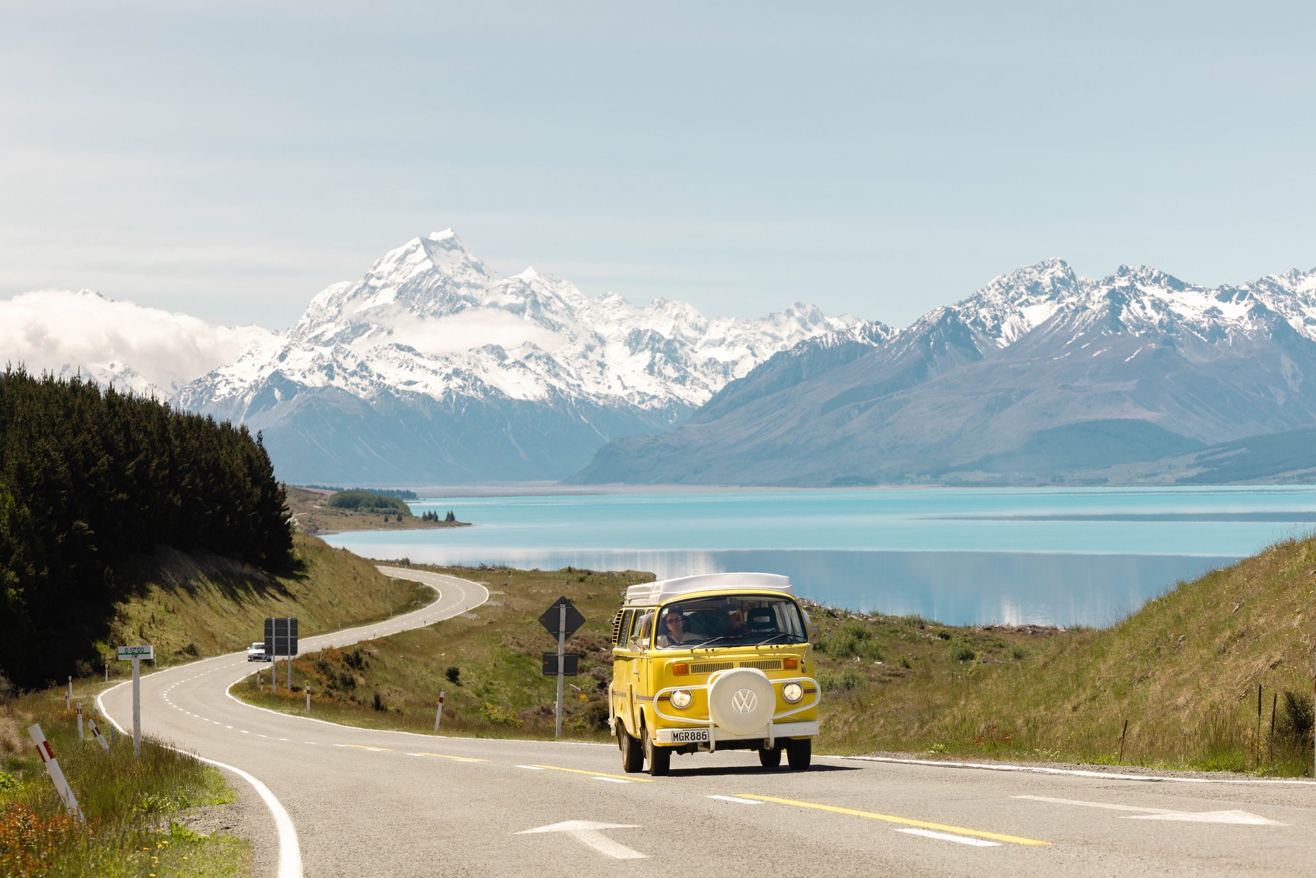 A yellow van drives on a winding road beside a turquoise lake, with snow-capped mountains in the background under a clear sky.
