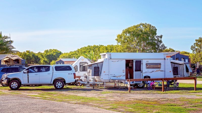A white SUV is parked next to a large caravan at a campsite, surrounded by trees and clear blue sky.