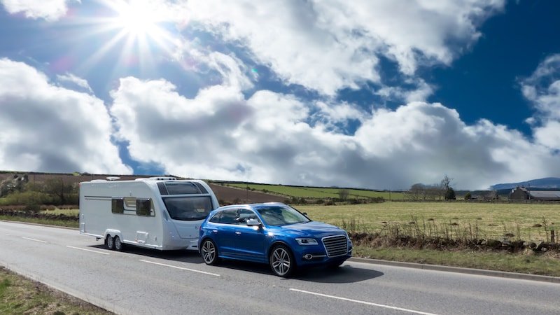 A blue SUV towing a white caravan on a rural road under a bright, sunny sky with scattered clouds and green fields in the background.